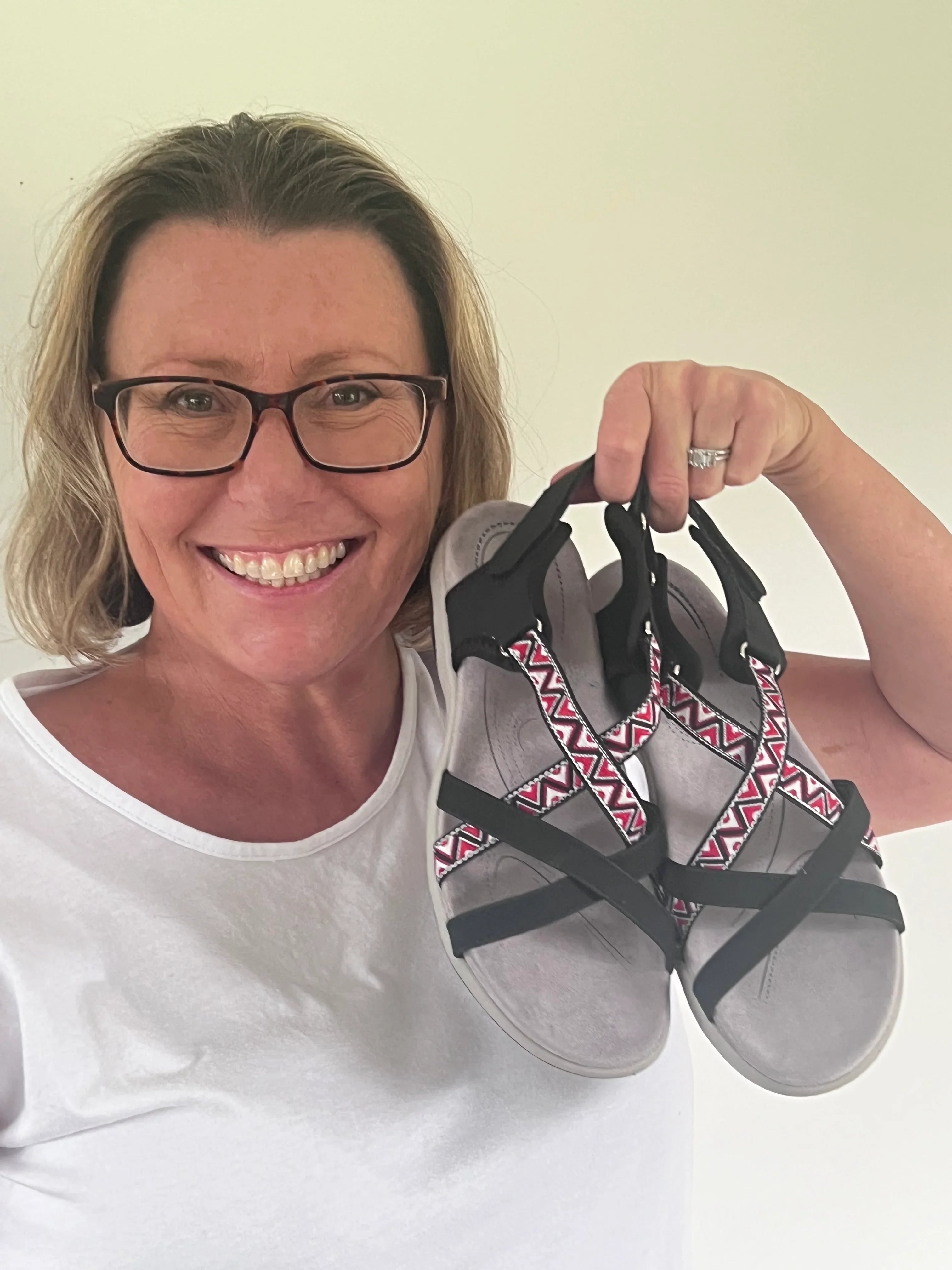 Smiling woman in glasses holding a pair of black sandals with geometric patterned straps
